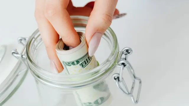 A close-up of a hand placing rolled dollars into a glass jar, symbolizing savings.