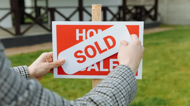 Close-up of hands placing a sold sticker on a real estate sign outside a house.