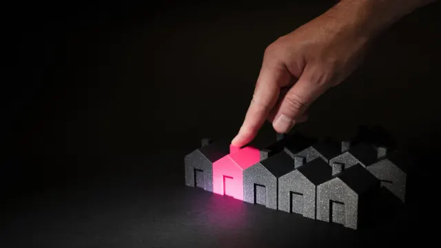A hand selecting a glowing pink house among dark miniature houses, symbolizing real estate choices.
